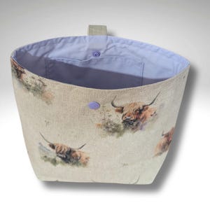 May include: A beige fabric tote bag with a repeating pattern of Highland cows in a watercolour style. The bag has a light blue interior, a small pocket, and a lavender button closure. A small loop is attached to the top.
