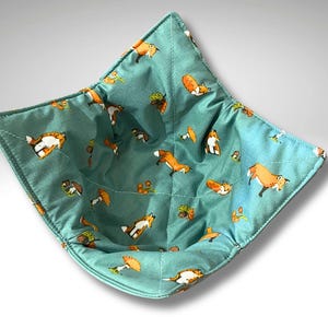 May include: A teal blue fabric bowl with a fox and mushroom print. The bowl is quilted and has a square shape.