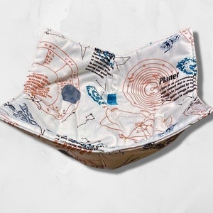 May include: A white fabric bowl with a celestial map design in shades of brown, blue, and black. The bowl features text and illustrations of planets and constellations. The bowl is designed to hold food.