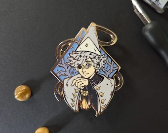 May include: A diamond-shaped enamel pin featuring a character with messy hair, a white pointed hat, and a monocle over one eye. The character wears a white coat with gold buttons over a dark shirt. The background is light blue with tree silhouettes.