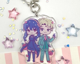 May include: A clear acrylic keychain featuring a cartoon couple. The female character has blue hair, a black dress, and red shoes. The male character has blonde hair, a green suit, and brown shoes. The keychain has a silver metal ring.