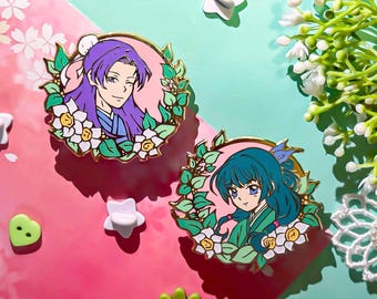Könnte beinhalten: Zwei Emaille-Pins mit Anime-Charakteren und floralen Details. Die Pins sind goldfarben mit rosa Hintergrund und zeigen grüne Blätter und weiße Blüten. Ein Pin zeigt einen Charakter mit lila Haaren, blauen Augen und einem blau-weißen Outfit. Der andere Pin zeigt einen Charakter mit blauen Haaren, blauen Augen und einem grün-weißen Outfit.