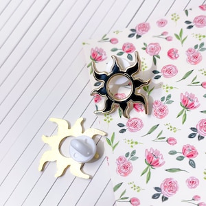 May include: Two gold sun pinback buttons. One sun is black with a gold outline. The other sun is gold. Both suns have a pointed, stylized design.