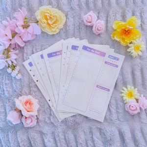 May include: A stack of white to-do list notepaper with purple accents, featuring the text "to-do list:" and "don't forget." The paper is surrounded by pink and yellow artificial flowers on a soft, gray textured surface.