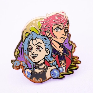 May include: A gold-colored enamel pin featuring two cartoon characters. The character on the left has blue hair and is wearing a black shirt. The character on the right has pink hair and is wearing a red shirt. The pin has a colorful background with a yellow moon.