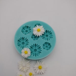 Puede incluir: Un molde de silicona turquesa con ocho cavidades en forma de margarita. Hay varias margaritas blancas con centros amarillos esparcidas alrededor del molde.