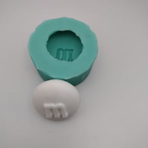 May include: A teal silicone mold with a round impression of an M&M candy. A white candy with an M on it is next to the mold.