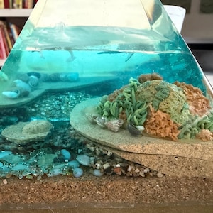 May include: A clear, pyramid-shaped resin art piece depicting an underwater scene. The layers include sand, pebbles, and a coral reef with green, orange, and beige elements. The top layer simulates water with small fish.