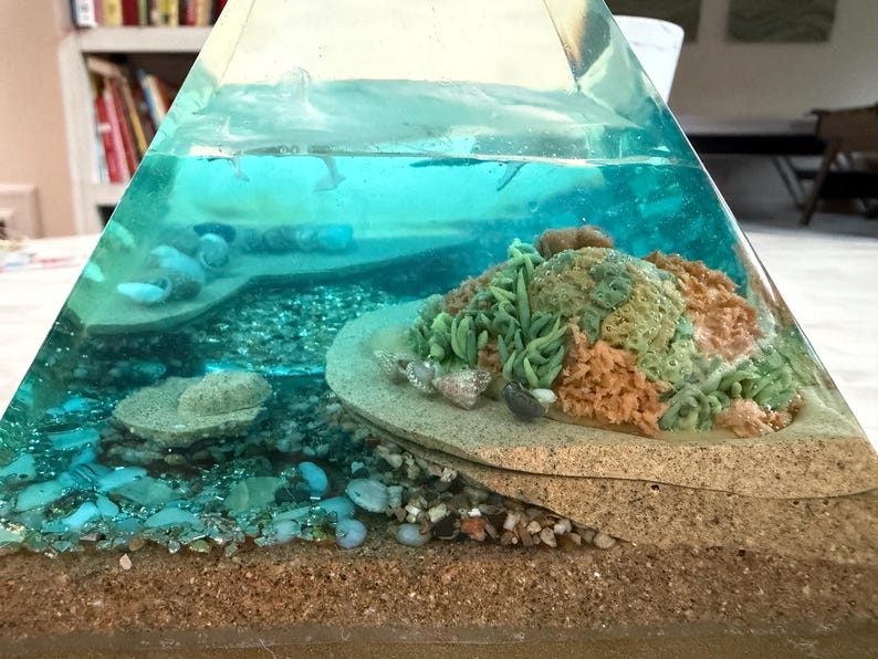 May include: A clear, pyramid-shaped resin art piece depicting an underwater scene. The layers include sand, pebbles, and a coral reef with green, orange, and beige elements. The top layer simulates water with small fish.