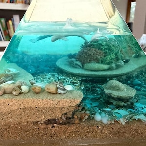 May include: A clear, pyramid-shaped resin art piece featuring an underwater scene. The scene includes a sandy seabed, coral, seashells, and small, gray dolphin figures swimming in the turquoise water. The base is a sandy color.