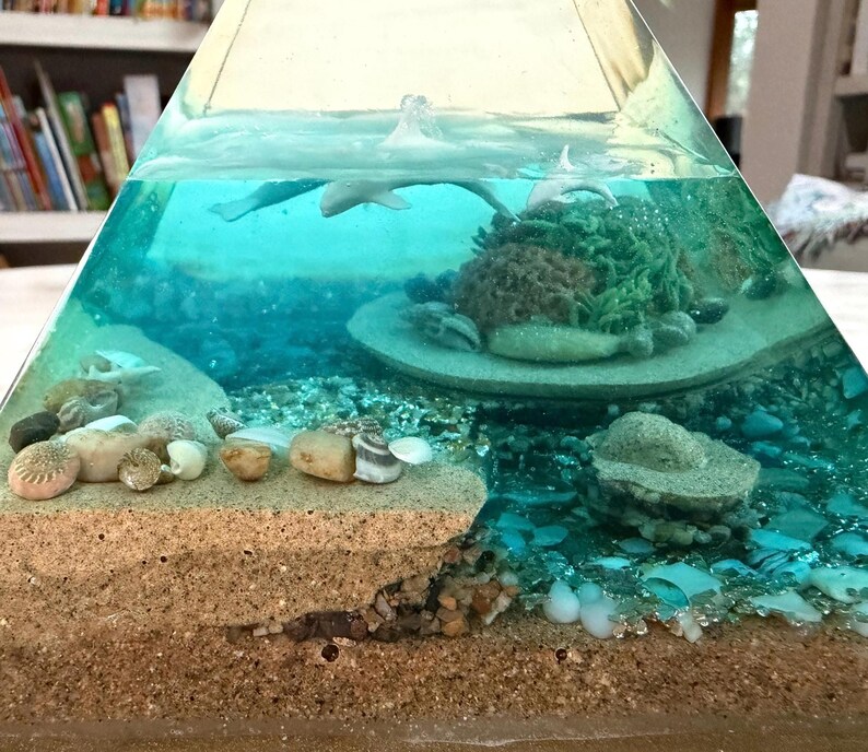May include: A clear, pyramid-shaped resin art piece featuring an underwater scene. The scene includes a sandy seabed, coral, seashells, and small, gray dolphin figures swimming in the turquoise water. The base is a sandy color.