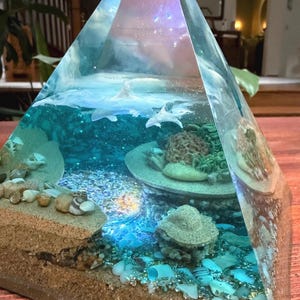 May include: A clear resin pyramid sculpture with an underwater scene. The sculpture features a sandy beach, ocean water, coral, and small sharks. The top of the pyramid has a galaxy design.