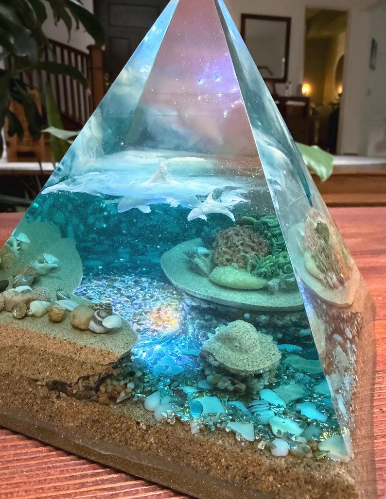 May include: A clear resin pyramid sculpture with an underwater scene. The sculpture features a sandy beach, ocean water, coral, and small sharks. The top of the pyramid has a galaxy design.