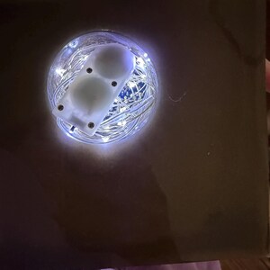 May include: A brown box with a circular opening revealing a glowing display. Inside the opening, a cluster of tiny white lights surrounds a white, rectangular device with two white, spherical objects. The device is secured with small screws.