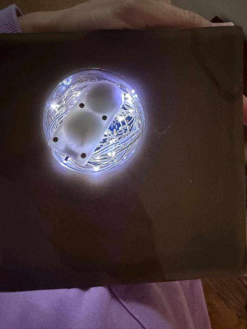 May include: A brown box with a circular opening revealing a glowing display. Inside the opening, a cluster of tiny white lights surrounds a white, rectangular device with two white, spherical objects. The device is secured with small screws.