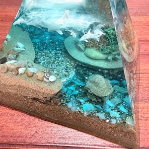 May include: A clear resin pyramid sculpture with a miniature ocean scene. The sculpture features sand, seashells, and small sharks. The ocean is depicted with blue and white resin, creating a realistic underwater effect. The base of the pyramid is brown.