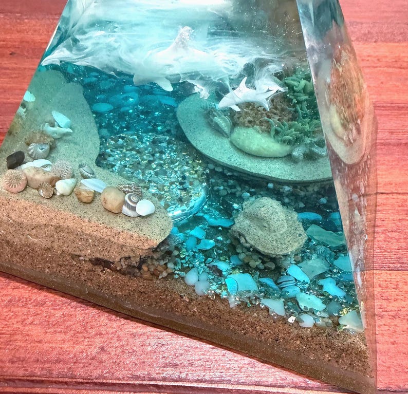 May include: A clear resin pyramid sculpture with a miniature ocean scene. The sculpture features sand, seashells, and small sharks. The ocean is depicted with blue and white resin, creating a realistic underwater effect. The base of the pyramid is brown.