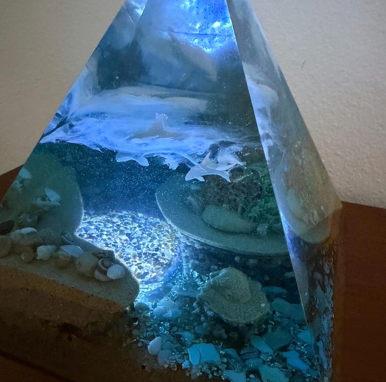 May include: A clear, pyramid-shaped resin sculpture with a seascape design. The interior features a miniature ocean scene with sand, rocks, and a shark. The resin is tinted blue, creating an underwater effect. The sculpture is illuminated from within.