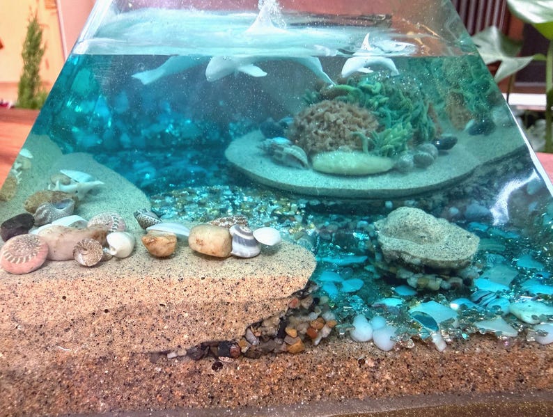 May include: A clear, pyramid-shaped decorative piece with a miniature ocean scene. It features a sandy base, seashells, and a turquoise-colored liquid with small, white dolphin figures. The scene includes a coral-like structure and small pebbles.