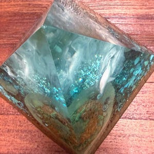 May include: A clear resin pyramid with a turquoise and white interior design. The pyramid contains a white, fish-like figure and other abstract shapes. The pyramid is set on a wooden surface.