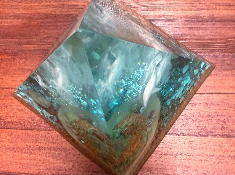 May include: A clear resin pyramid with a turquoise and white interior design. The pyramid contains a white, fish-like figure and other abstract shapes. The pyramid is set on a wooden surface.