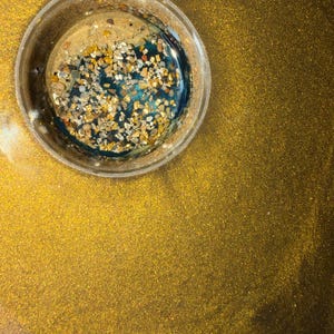 May include: A clear, circular resin piece with a gold glitter background. The resin contains gold and tan-colored stones and a deep teal-colored liquid. The background is a solid gold color with glitter.