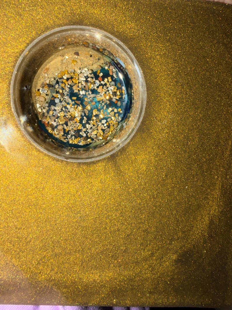 May include: A clear, circular resin piece with a gold glitter background. The resin contains gold and tan-colored stones and a deep teal-colored liquid. The background is a solid gold color with glitter.