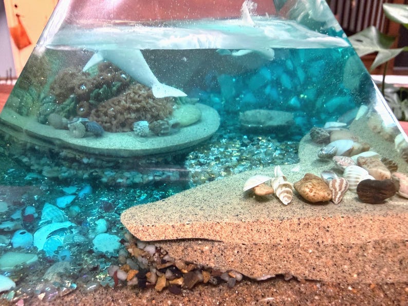 May include: A clear, pyramid-shaped resin art piece with a miniature ocean scene. It features a white dolphin, coral, seashells, and sand. The water is a vibrant blue, and the base is a sandy color, creating a layered effect.