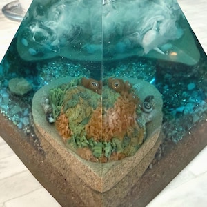 May include: A pyramid-shaped resin art piece featuring an underwater scene. The artwork showcases a heart-shaped coral reef with green and orange hues, surrounded by blue resin, small stones, and white accents, creating a marine environment.