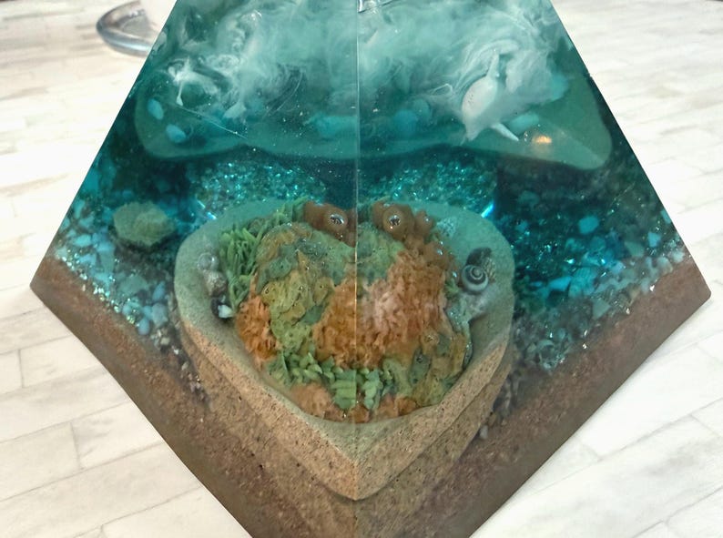 May include: A pyramid-shaped resin art piece featuring an underwater scene. The artwork showcases a heart-shaped coral reef with green and orange hues, surrounded by blue resin, small stones, and white accents, creating a marine environment.
