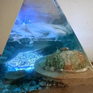 May include: A clear resin pyramid sculpture with an underwater scene. The sculpture features a sandy base, rocks, and a coral reef. The water is a mix of blue and white, with a blue light shining through the top.