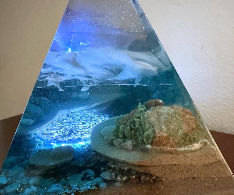 May include: A clear resin pyramid sculpture with an underwater scene. The sculpture features a sandy base, rocks, and a coral reef. The water is a mix of blue and white, with a blue light shining through the top.