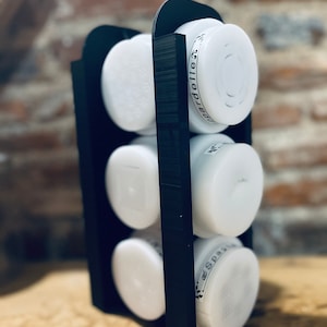 Peut inclure: Un porte-épices en plastique noir avec six pots à épices ronds blancs. Les pots ont des étiquettes noires avec du texte blanc. Les étiquettes indiquent "Gardelle" et "Spas".