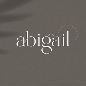 May include: The name "abigail" is displayed in a white, elegant font on a muted gray background. The word is stylized with a unique ligature, and the phrase "unique ligature font" is arched above the name.