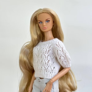Puede incluir: Una muñeca de moda con cabello largo rubio y pecas que viste un suéter de punto blanco de manga corta con un delicado patrón calado y pantalones de color gris claro. La muñeca tiene ojos azules, delineador oscuro y una expresión neutra.