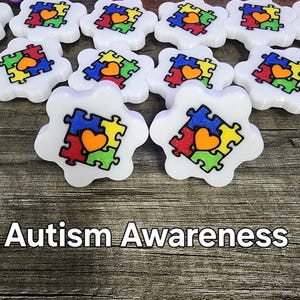 May include: White puzzle piece shaped charms with a colorful puzzle piece design and a red heart in the center. The charms say "Autism Awareness" at the bottom of the image.