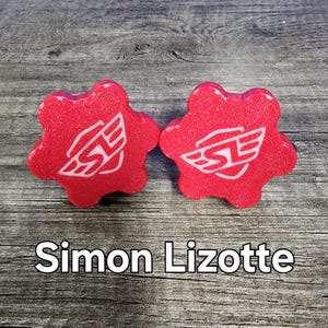May include: Two red, star-shaped, plastic gear-shaped knobs with a white logo that says "SL" with wings. The logo is on a white background. The knobs are on a wooden surface. The text "Simon Lizotte" is below the knobs.