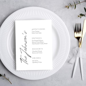 Może przedstawiać: Biała karta menu z napisem "The Johnson's" w eleganckim skrypcie, umieszczona na białym talerzu ze złotym widelcem i nożem. Menu zawiera przystawki, dania główne, desery i napoje.