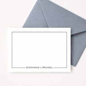 Puede incluir: Una tarjeta rectangular blanca con un borde negro fino y el texto "STEPHANIE + MICHAEL" impreso en negro en la parte inferior, acompañada de un sobre azul pizarra colocado detrás de la tarjeta sobre una superficie blanca.