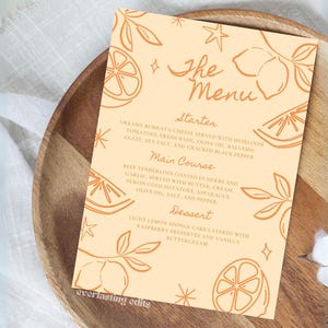 Puede incluir: Una tarjeta de menú color melocotón con ilustraciones de limones y el texto "The Menu". La tarjeta está colocada sobre un plato de madera. El menú enumera entrantes, platos principales y postres.