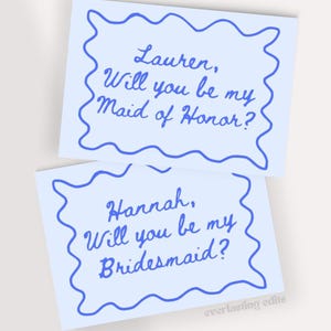 Könnte beinhalten: Zwei hellblaue rechteckige Karten mit einem wellenförmigen dunkelblauen Rand. Die obere Karte zeigt den Text, Lauren, Will you be my Maid of Honor? Die untere Karte zeigt, Hannah, Will you be my Bridesmaid? Beide in dunkelblauer Schreibschrift.