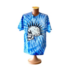 May include: A blue tie-dye t-shirt with a white skull graphic and a mohawk. The skull has black eyes and a black mouth.