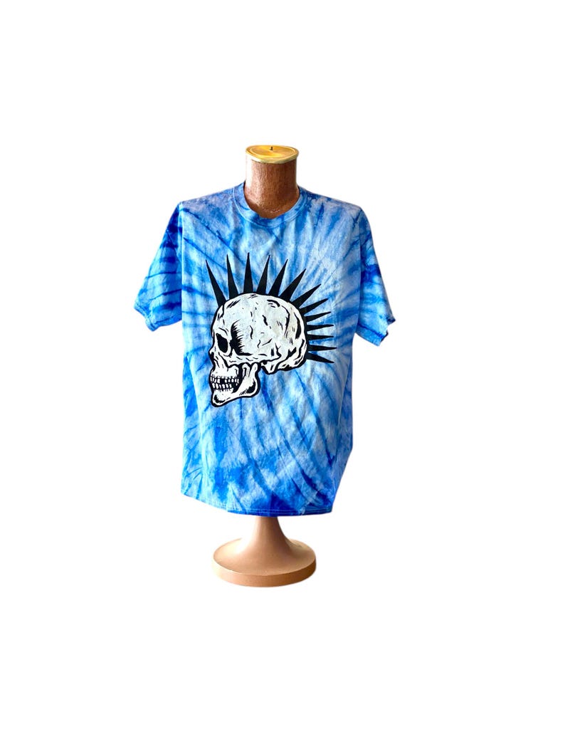 May include: A blue tie-dye t-shirt with a white skull graphic and a mohawk. The skull has black eyes and a black mouth.
