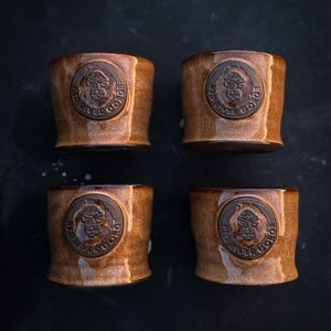 May include: Four brown ceramic cups with a textured finish. Each cup features a circular emblem with the text "Red River Gorge" and a detailed illustration. The cups are arranged on a dark surface.