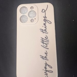 May include: A white phone case with a glittery camera lens cover and the text "enjoy the little things" written in black cursive.
