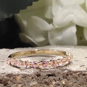 May include: A delicate gold band ring with a row of small pink gemstones. The ring is set against a background of white flowers.