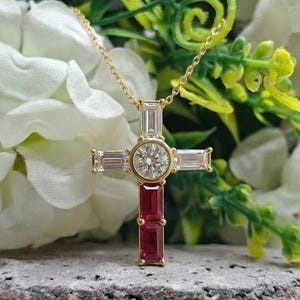 Pode incluir: Um colar com pingente de cruz dourada com uma corrente delicada. A cruz apresenta um diamante redondo central, diamantes lapidados em baguete e duas pedras preciosas vermelhas retangulares. O colar é exibido em um fundo de flores brancas e folhagem verde.