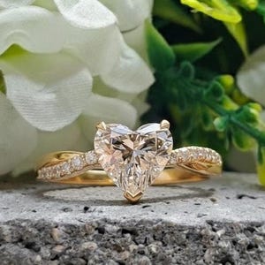 Può includere: Un anello di fidanzamento in oro con una pietra centrale a forma di cuore. L'anello presenta una fascia attorcigliata con piccoli diamanti di contorno. L'anello è posto su uno sfondo di fiori bianchi e fogliame verde.