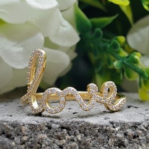 May include: A gold-colored ring spelling out the word "love" in cursive script. The ring is encrusted with small, clear gemstones. The ring is set against a textured gray surface, with white flowers and green foliage in the background.