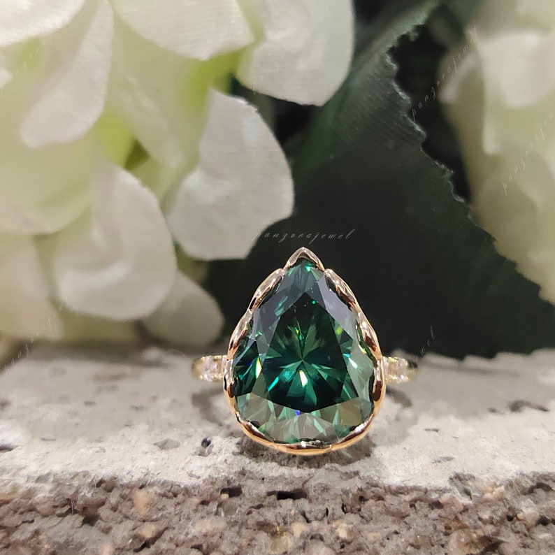 May include: A pear-shaped green gemstone ring with a gold band and a delicate, intricate design. The ring is set on a textured surface.